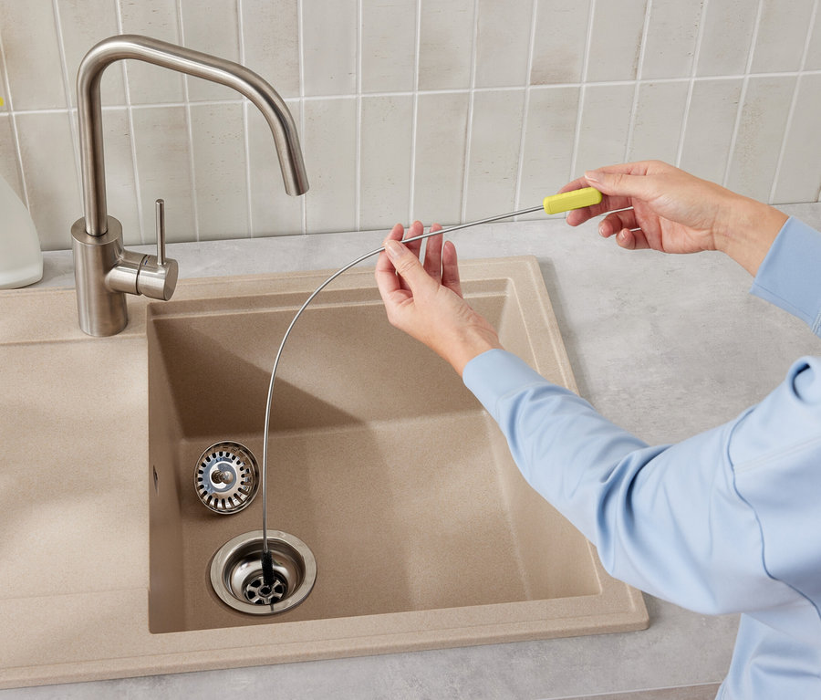 Kobieta czyści odpływ zlewu za pomocą narzędzia do czyszczenia odpływów z żółtą rączką.