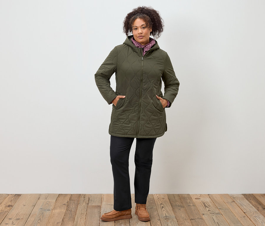 Kobieta stoi z rękami w kieszeniach oliwkowozielonej kurtki puchowej z kapturem, czarnych spodniach i brązowych mokasynach.