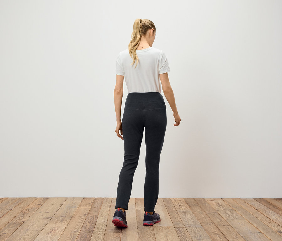 Kobieta z blond włosami związanymi w kucyk stoi tyłem do kamery, ubrana w czarne spodnie i białą koszulkę.