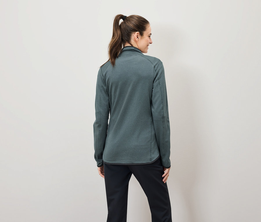 Z tyłu widać kobietę z brązowymi włosami związanymi w kucyk, ubraną w zieloną bluzę dresową zapinaną na zamek i czarne spodnie.