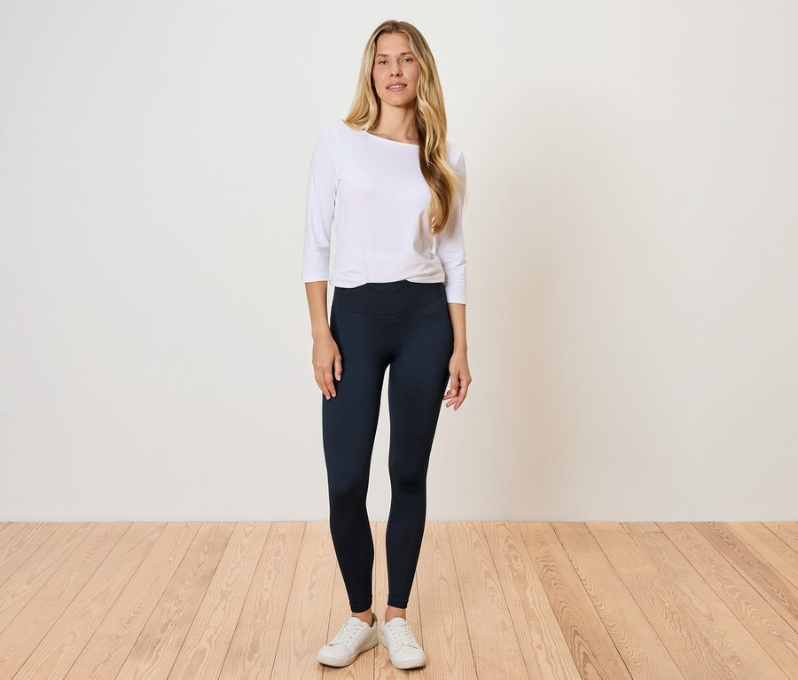 Kobieta stoi na drewnianej podłodze, ubrana w białą koszulę z długim rękawem i czarne legginsy.