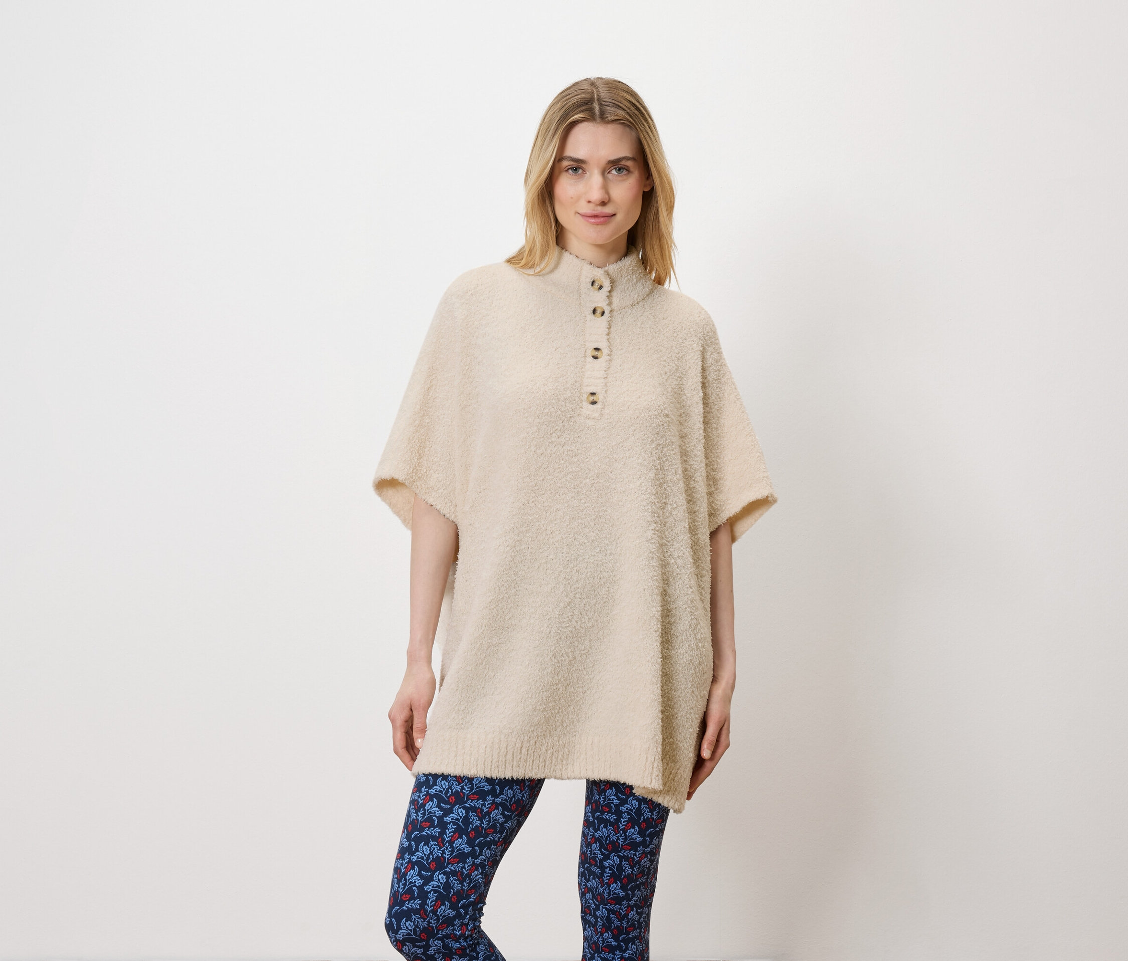 Kobieta pozuje w studio na białym tle. Ma na sobie beżowy sweter i niebieskie legginsy.