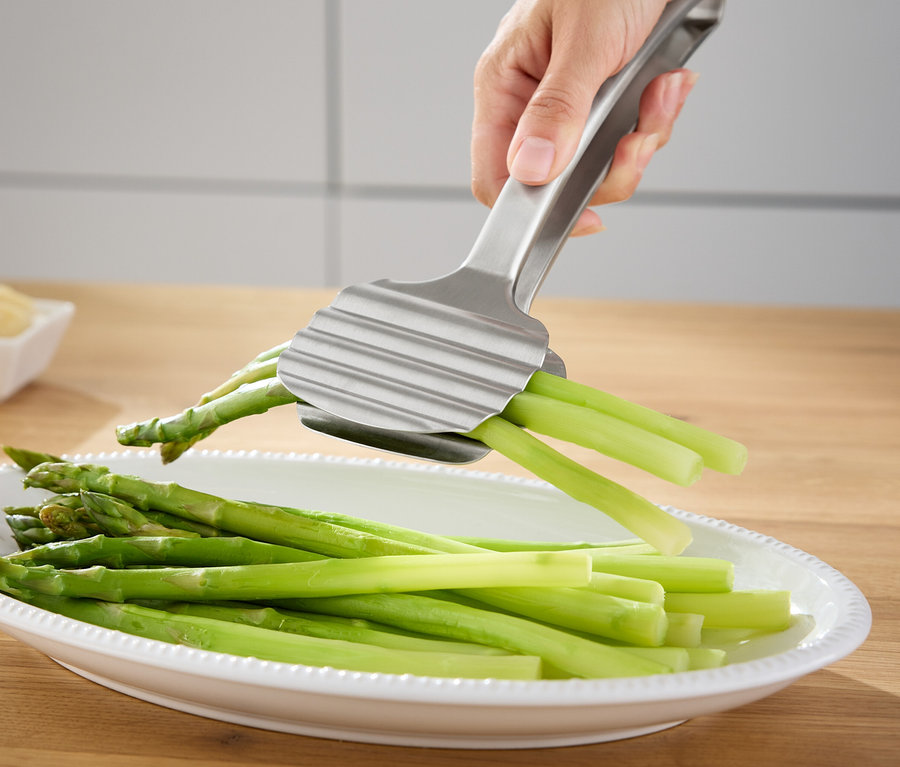Dłoń trzyma srebrne szczypce do szparagów nad talerzem z zielonymi szparagami.