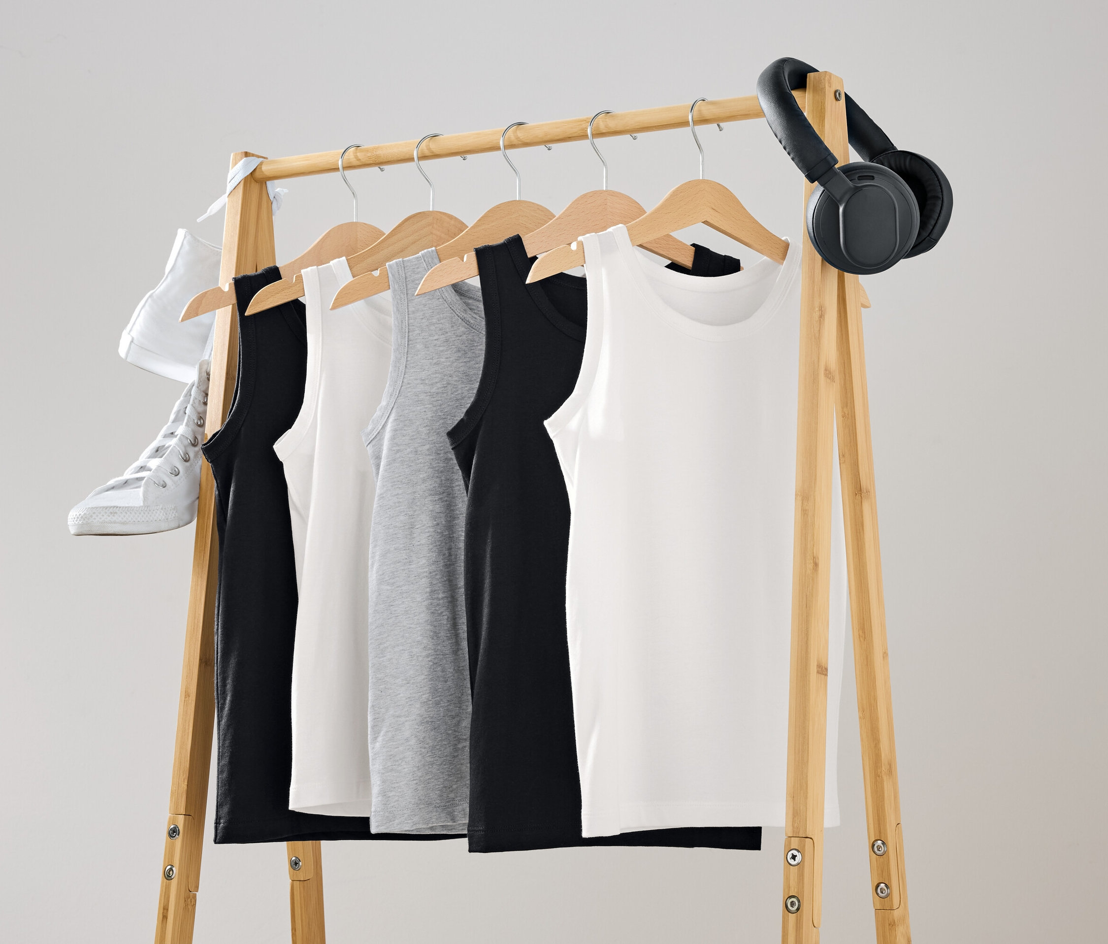 Na drewnianym wieszaku wiszą czarne, białe i szare podkoszulki. Na wieszaku zawieszony jest biały but i czarne słuchawki.