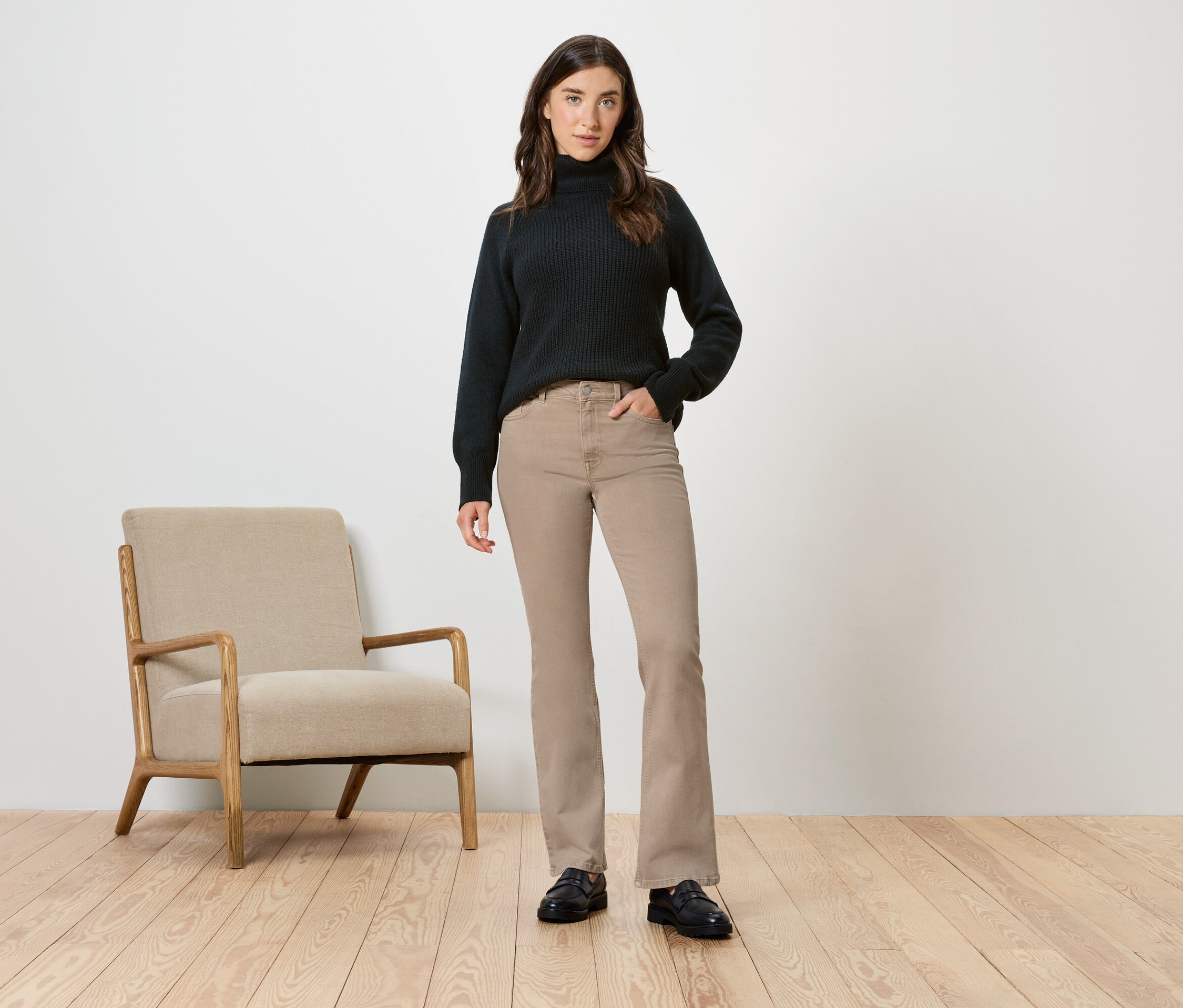 Kobieta w czarnym golfie i beżowych spodniach pozuje obok beżowego krzesła.