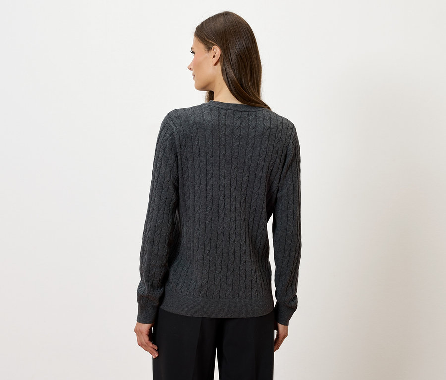 Kobieta stoi tyłem, ubrana w szary sweter z dzianiny w warkocze i czarne spodnie.