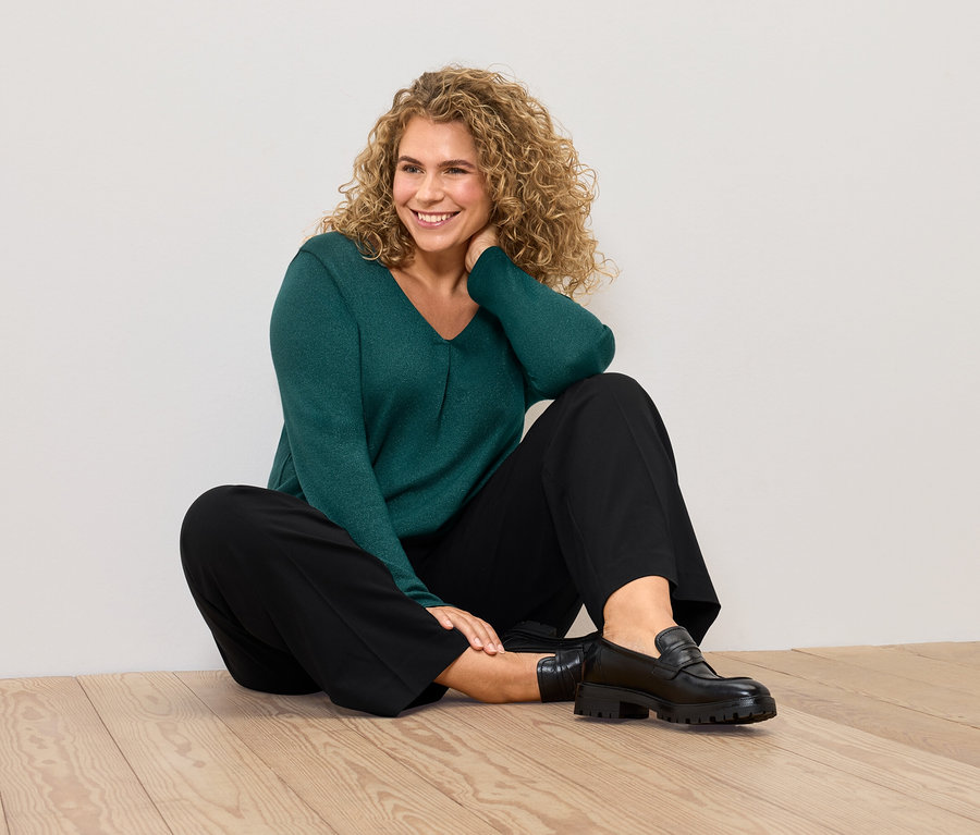Kobieta siedzi na podłodze w zielonym swetrze i czarnych spodniach.