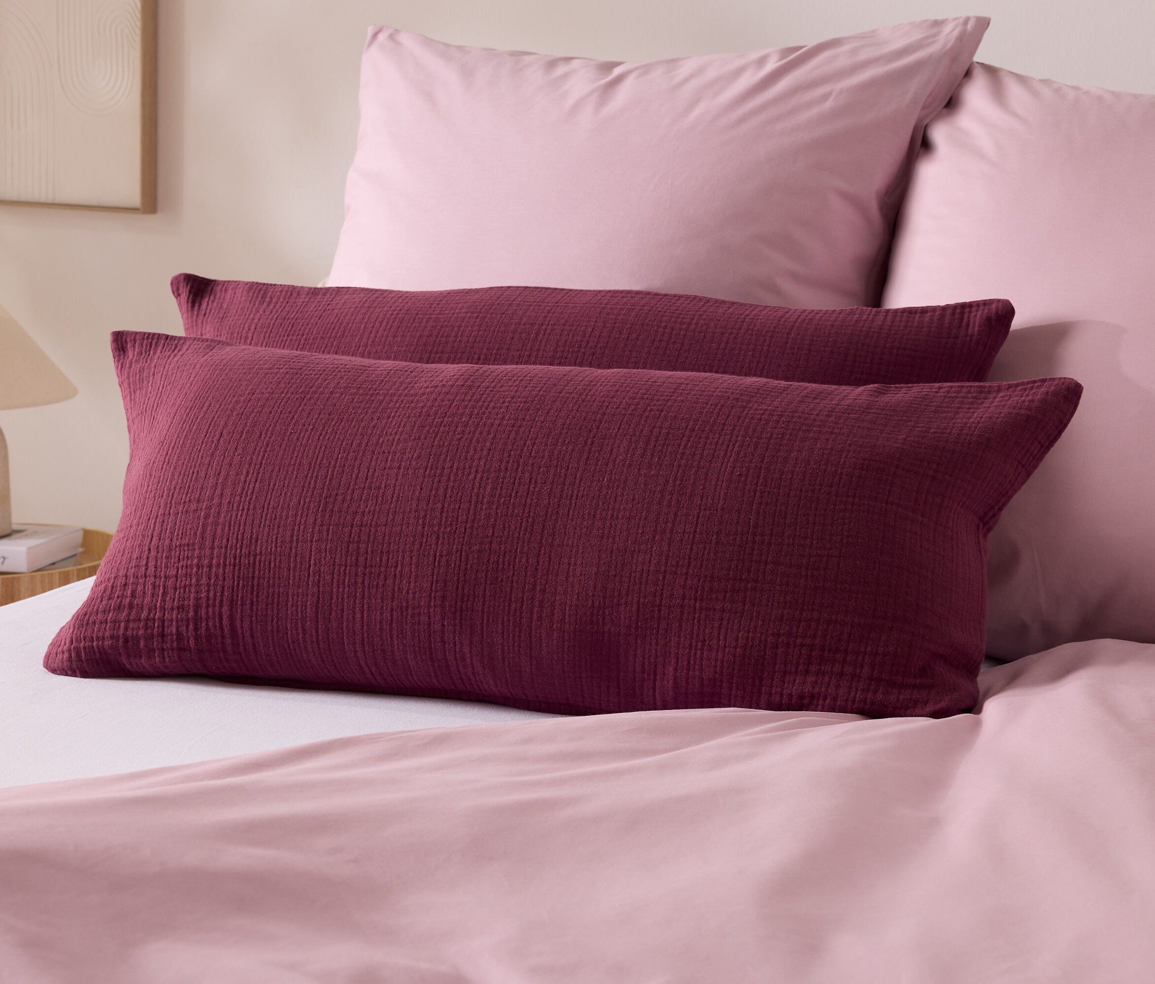 Dwie bordowe poduszki leżą na łóżku z różową pościelą. W tle dwie różowe poduszki i lampa.