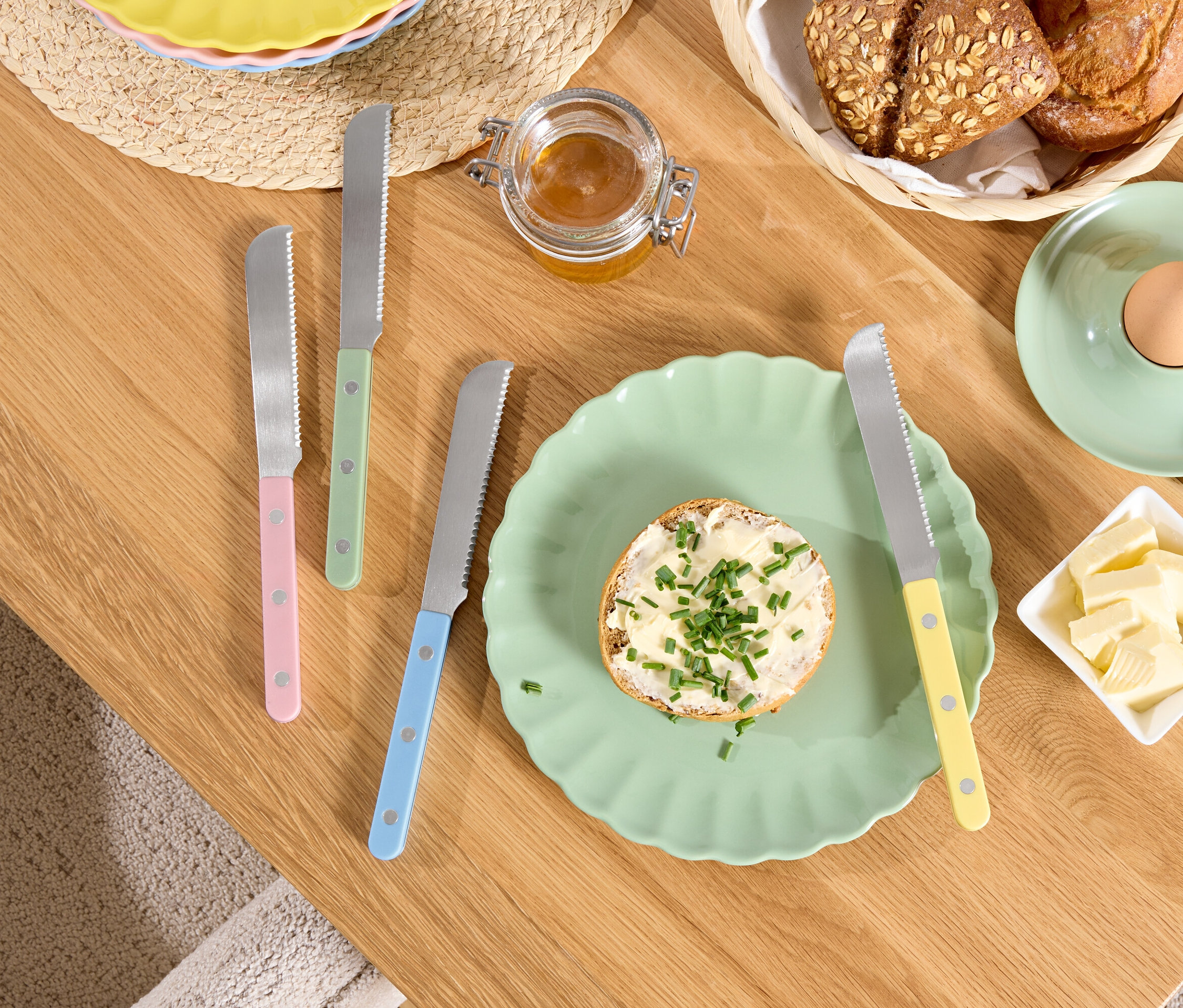 Na drewnianym stole leżą talerz z chlebem z masłem i ziołami, masło w misce, jajko w kieliszku i noże z kolorowymi rączkami.