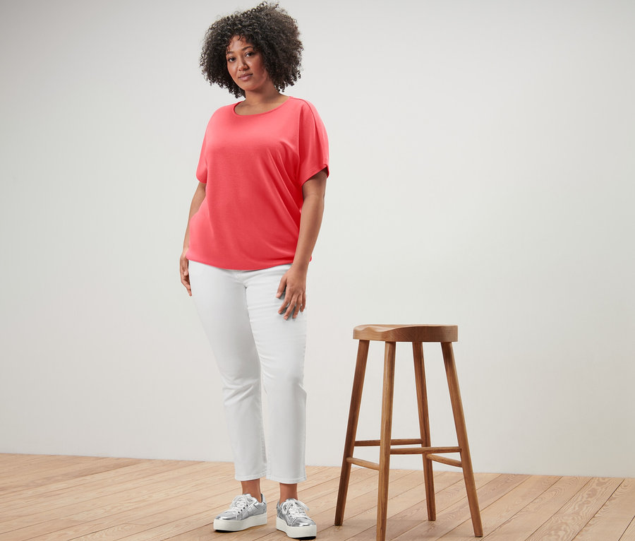 Kobieta stoi obok drewnianego stołka i ma na sobie koralową bluzkę.