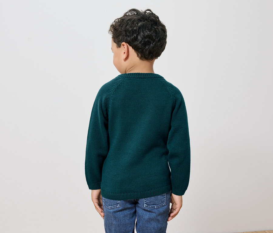 Chłopiec o ciemnych, kręconych włosach stoi tyłem do kamery, ubrany w zielony sweter i dżinsy.