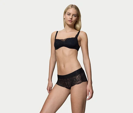 Blondynka stoi, ubrana w czarny koronkowy biustonosz i czarne koronkowe figi Triumph Body Makeup Illusion.