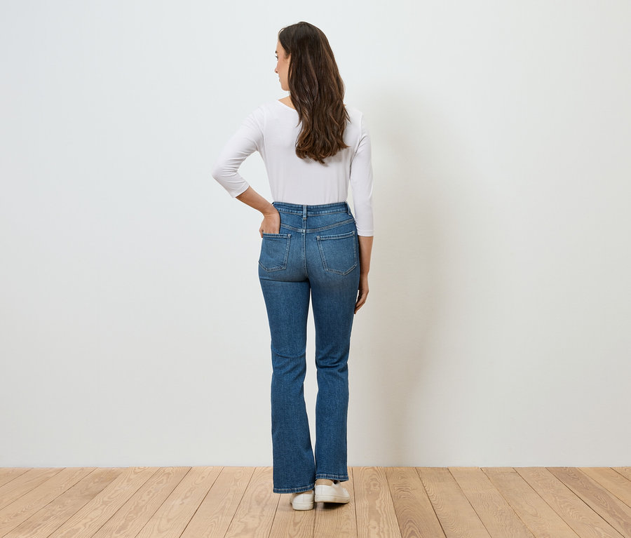 Kobieta tyłem do kamery trzyma rękę w kieszeni niebieskich dżinsów i ma na sobie białą koszulkę z długim rękawem.
