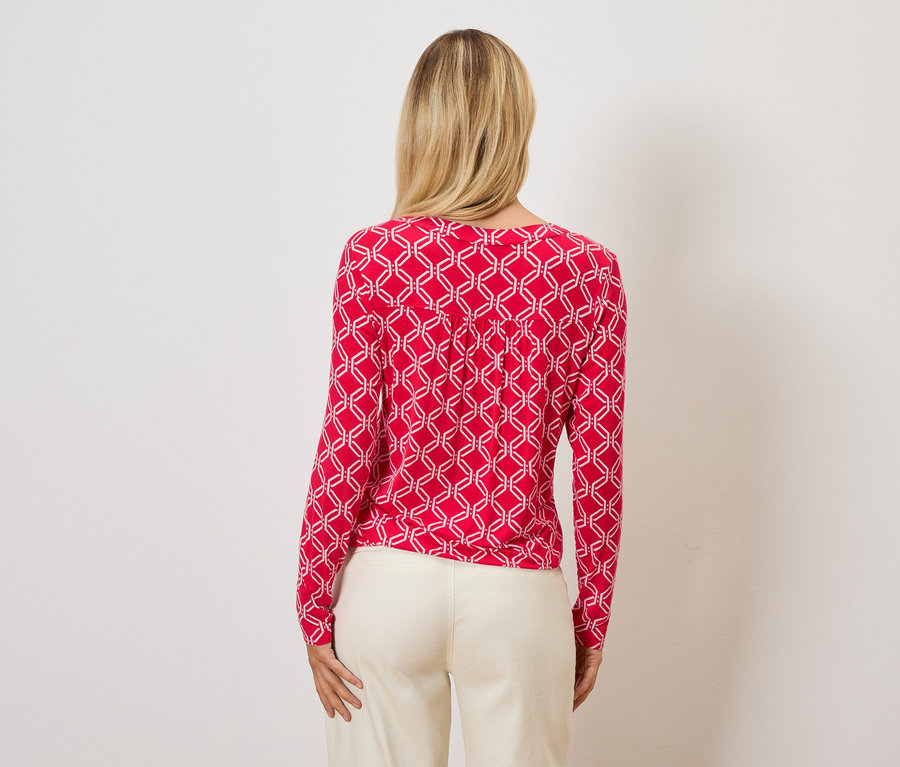 Z tyłu widać kobietę z blond włosami, ubraną w czerwoną bluzkę z białym geometrycznym wzorem i kremowe spodnie.