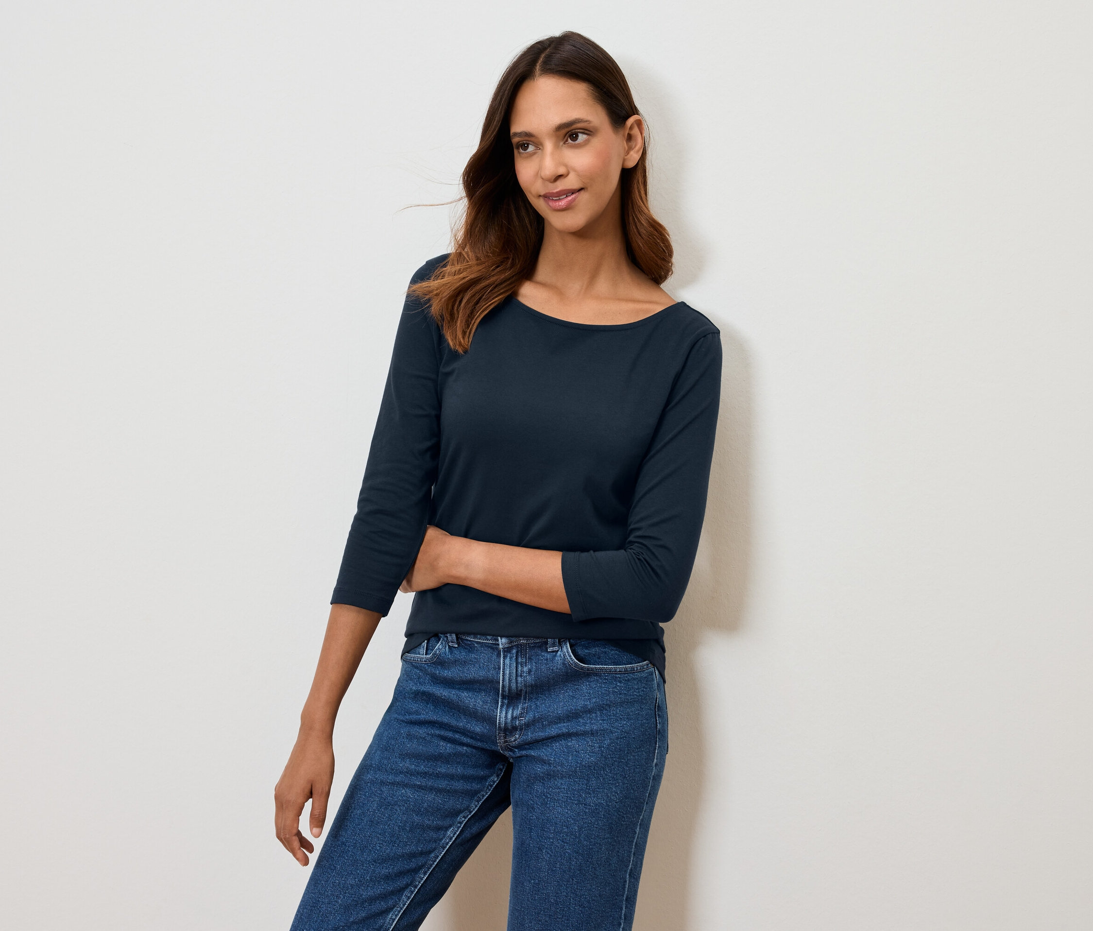 Kobieta pozuje w granatowej koszulce z rękawem trzy czwarte i niebieskich jeansach.