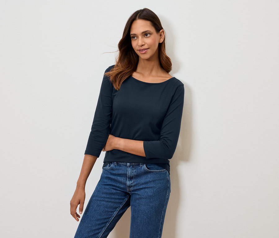 Kobieta pozuje w granatowej koszulce z rękawem trzy czwarte i niebieskich jeansach.