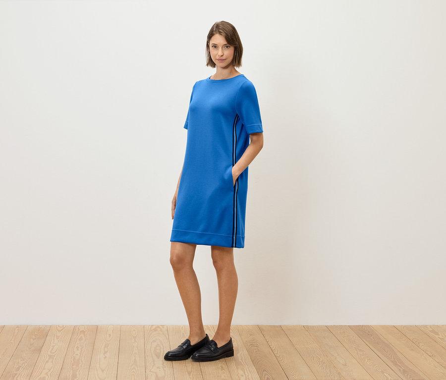 Kobieta stoi i pozuje w niebieskiej sukience z krótkim rękawem i czarnych mokasynach.