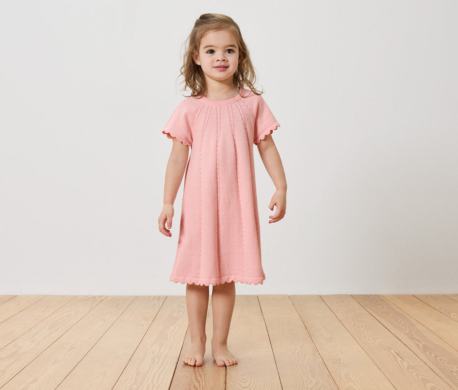 Dziewczynka stoi boso na drewnianej podłodze, ubrana w różową sukienkę.