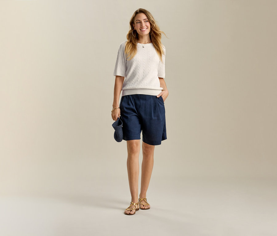 Uśmiechnięta kobieta w jasnej bluzce i granatowych szortach Leinen-Bermuda-Shorts, z ręką w kieszeni.