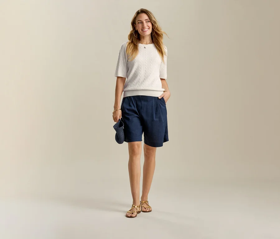 Uśmiechnięta kobieta w jasnej bluzce i granatowych szortach Leinen-Bermuda-Shorts, z ręką w kieszeni.