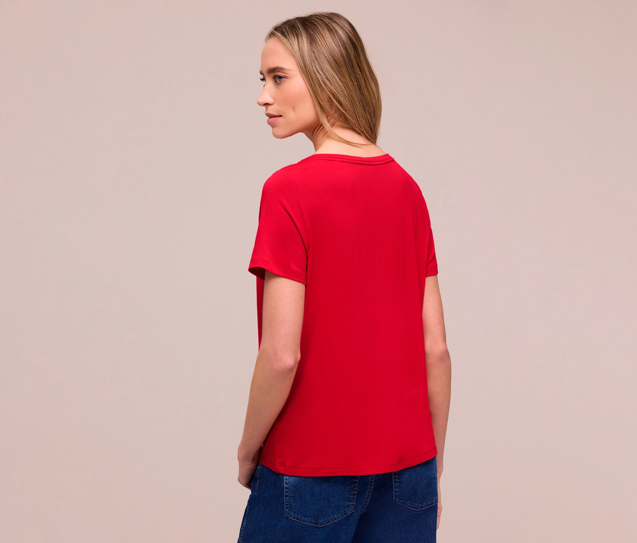 Kobieta od tyłu w czerwonej koszulce Street One i dżinsach. Patrzy w lewo.