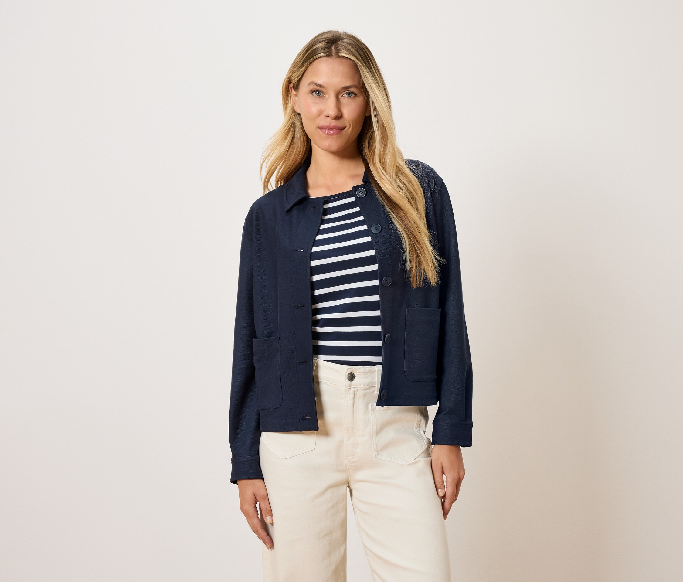 Kobieta ma na sobie granatową kurtkę nałożoną na bluzkę w paski i kremowe spodnie.