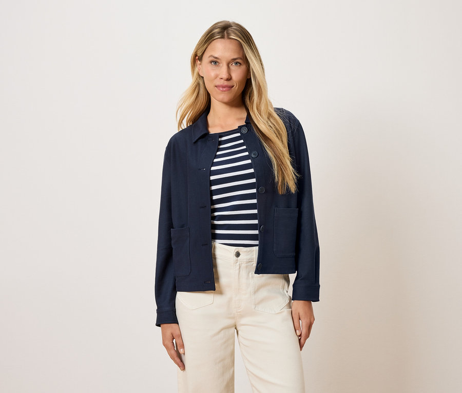 Kobieta ma na sobie granatową kurtkę nałożoną na bluzkę w paski i kremowe spodnie.