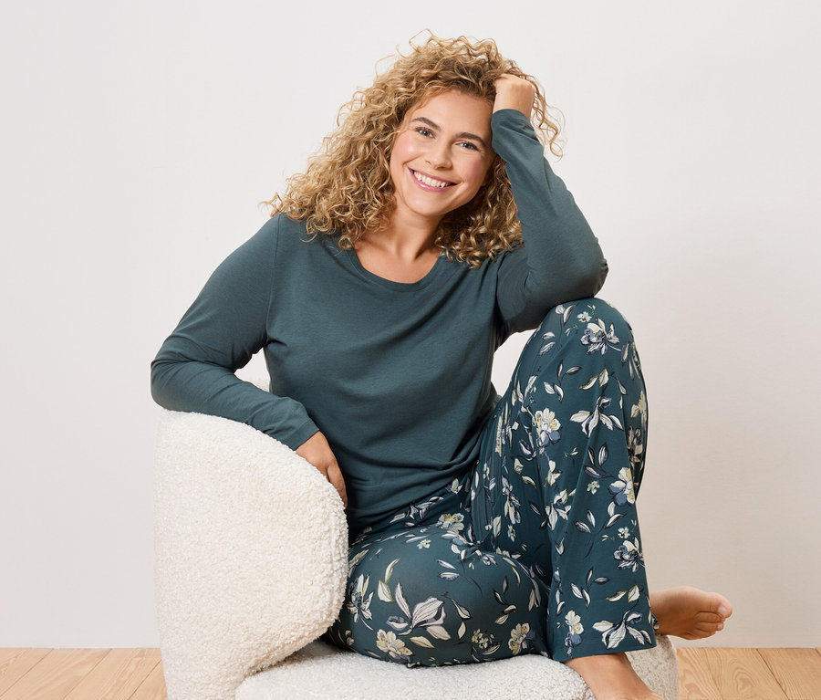 Kobieta z kręconymi włosami siedzi w fotelu, ubrana w ciemnozieloną piżamę w kwiaty.