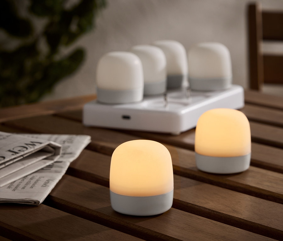 Dwie podświetlane, bezprzewodowe lampy stołowe na drewnianym stole, ze stacją ładującą i czterema dodatkowymi lampami w tle. Obok leży gazeta.