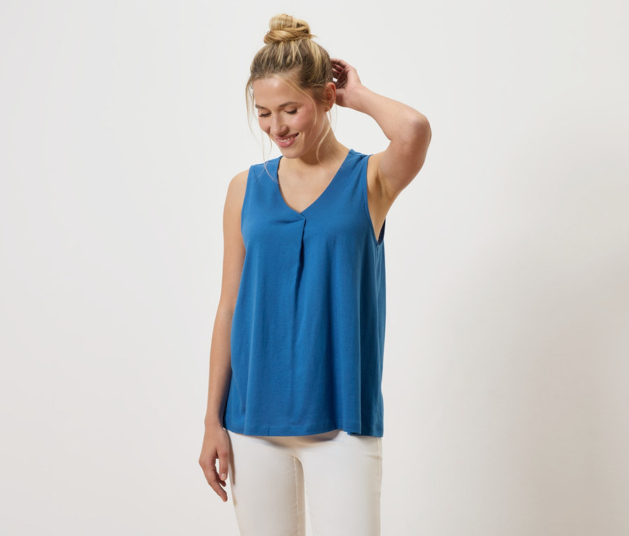 Kobieta z blond włosami ma na sobie niebieski top z dekoltem w serek i białe spodnie.