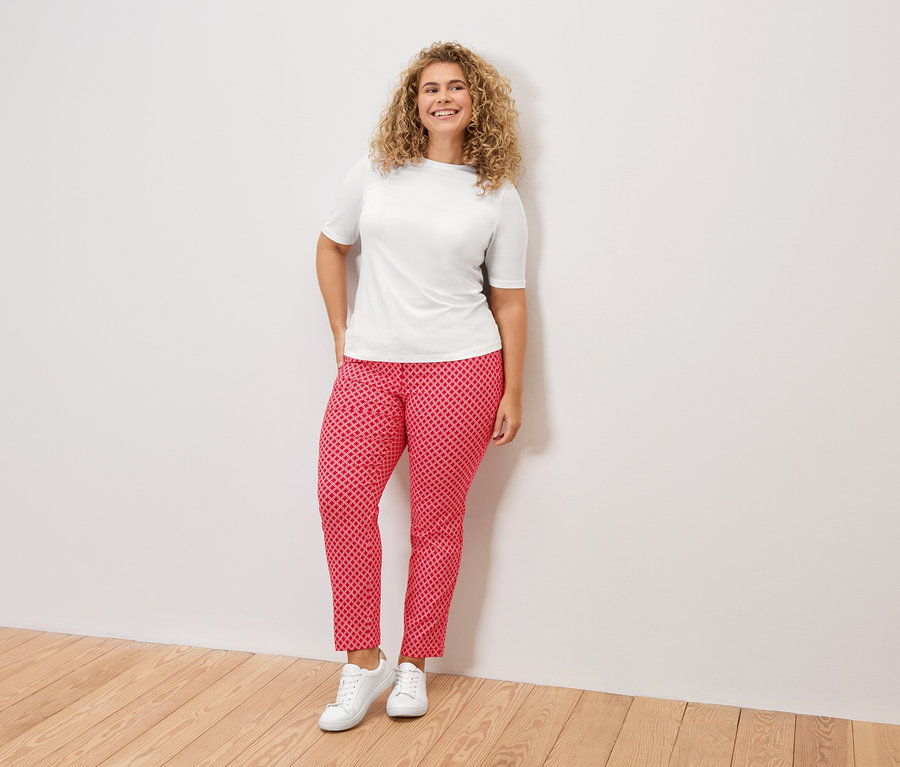 Kobieta z kręconymi włosami w białej koszulce, czerwonych wzorzystych spodniach i białych tenisówkach stoi na tle białej ściany.