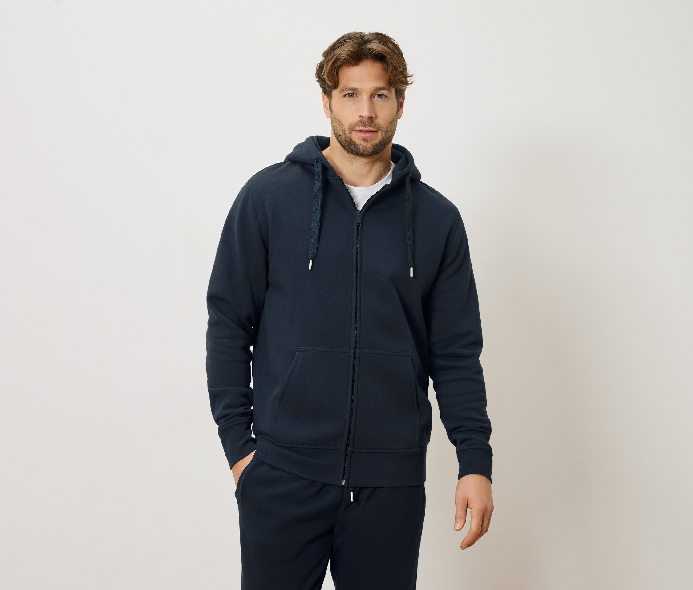 Mężczyzna w granatowej bluzie z kapturem na zamek błyskawiczny i granatowych spodniach dresowych, stojący na kremowym tle.