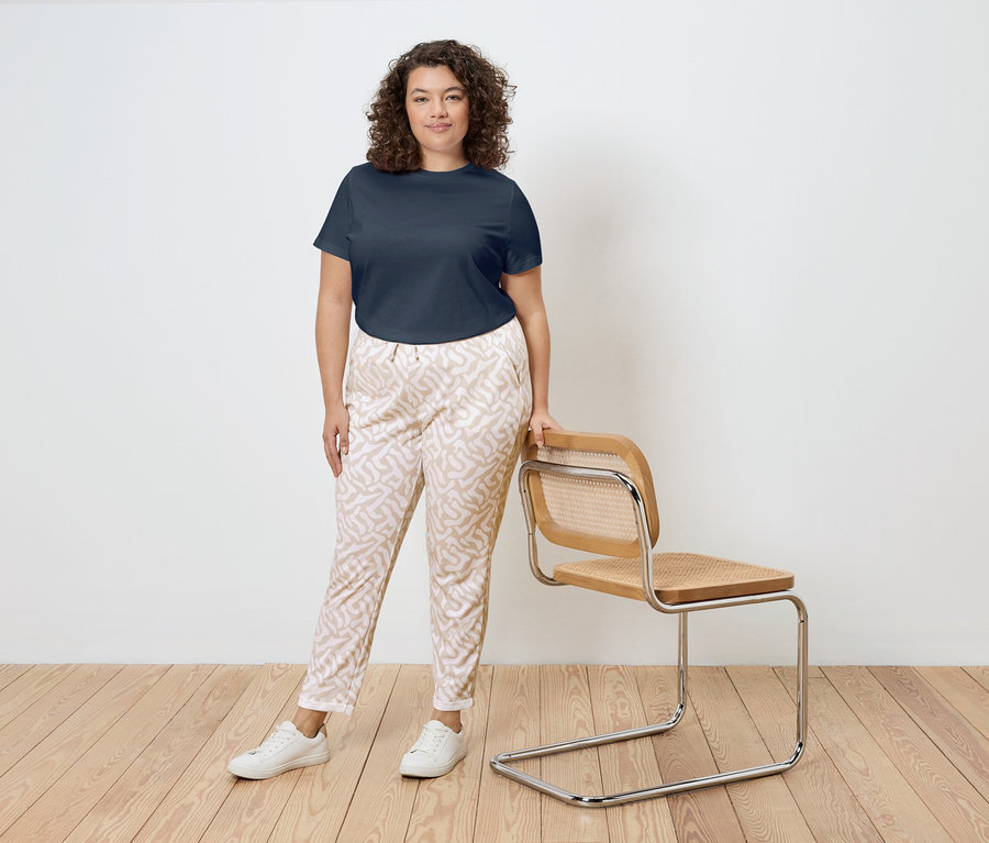 Kobieta o kręconych włosach stoi obok krzesła z drewnianą ramą i rattanowym siedziskiem. Ma na sobie granatowy T-shirt, wzorzyste spodnie i białe tenisówki.