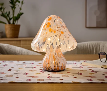 Grzybowa lampa z pomarańczowo-białego nakrapianego szkła z wewnętrznymi światłami stoi na drewnianym stole. Pod nią leży biały bieżnik z wzorami grzybów. Po prawej stronie leży otwarta książka z okularami w ciemnych oprawkach. W rozmytym tle widoczna jest roślina doniczkowa i oprawiony obraz.