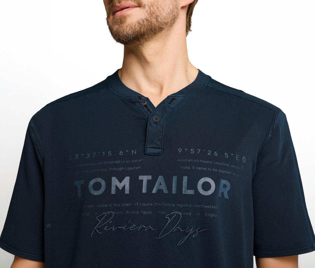 Mężczyzna w granatowej koszulce Henley z nadrukiem 'TOM TAILOR', 'Riviera Days' i współrzędnymi geograficznymi.