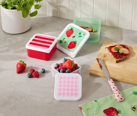 Na kuchennym blacie stoi kilka kolorowych pojemników na żywność, niektóre wypełnione owocami, obok kromki chleba i noża.