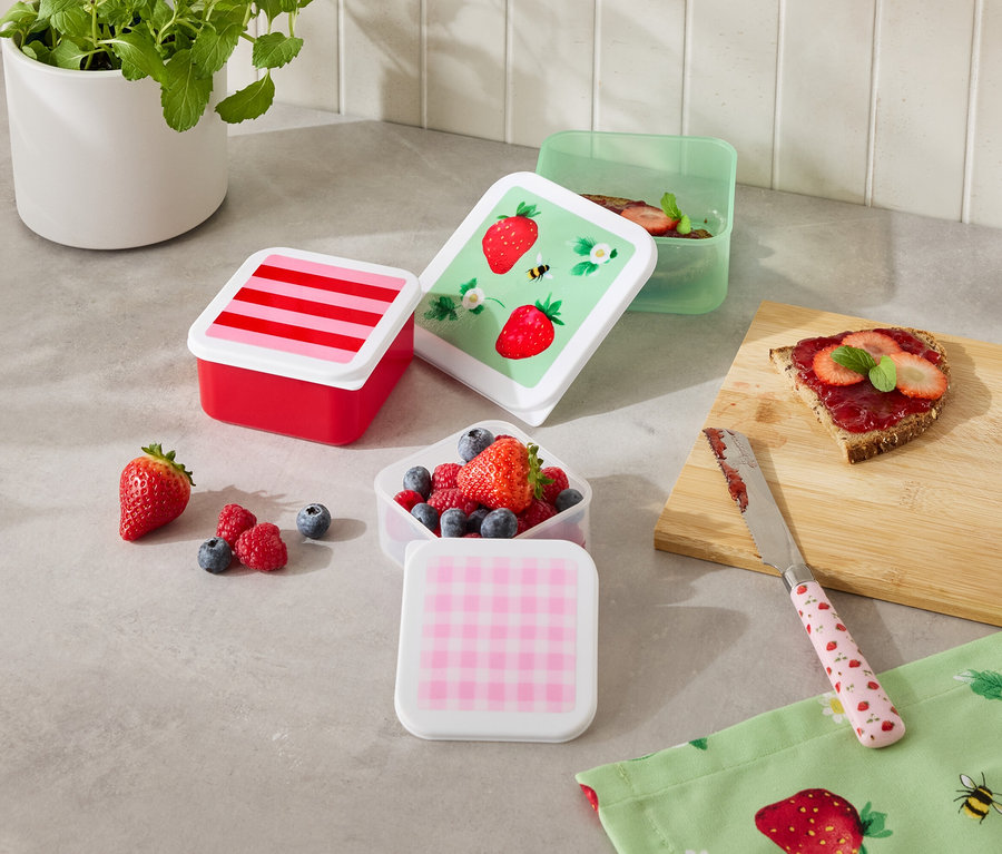 Na kuchennym blacie stoi kilka kolorowych pojemników na żywność, niektóre wypełnione owocami, obok kromki chleba i noża.
