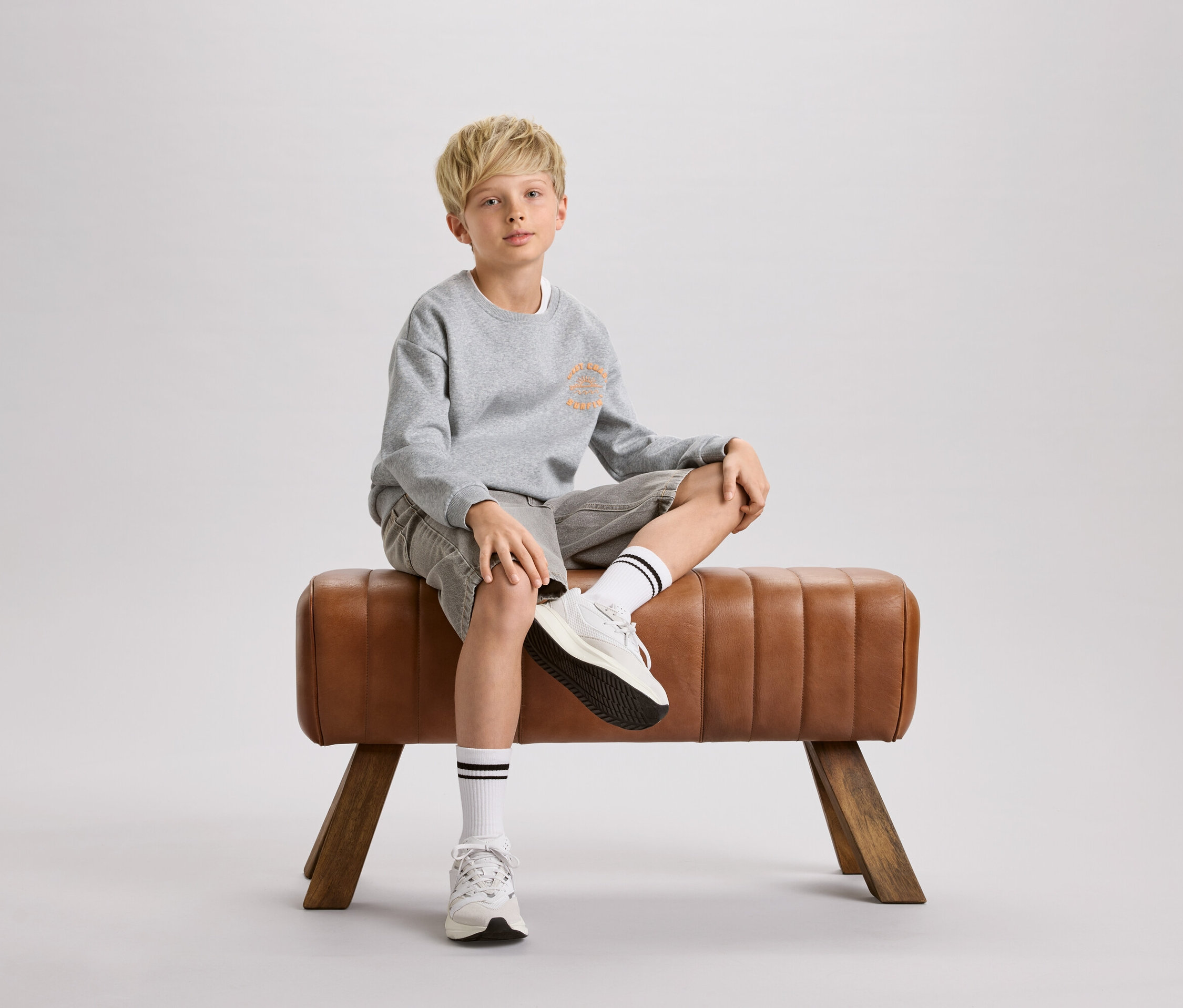 Chłopiec o blond włosach siedzi na brązowym kozle gimnastycznym. Ma na sobie szarą bluzę, szorty i białe skarpetki z czarnymi paskami.