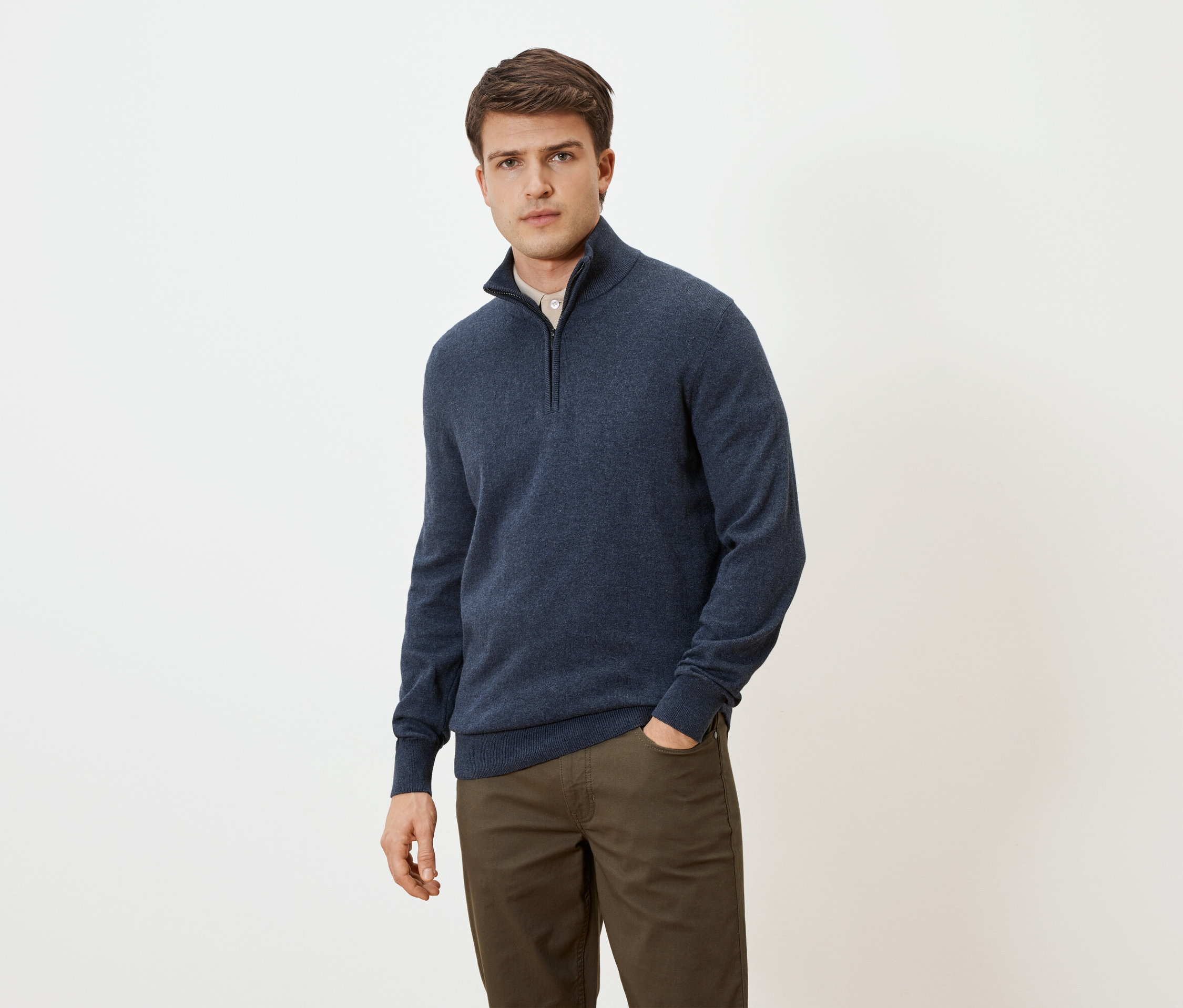 Mężczyzna w niebieskim swetrze na zamek i brązowych spodniach, z ręką w kieszeni, na neutralnym tle.