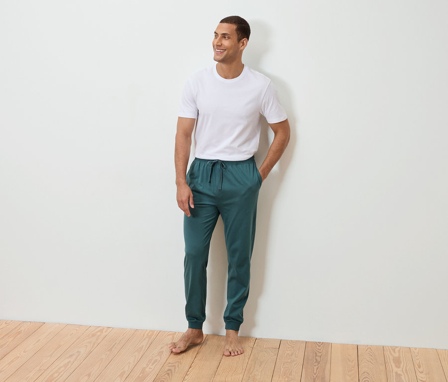 Mężczyzna stoi boso z ręką w kieszeni na tle białej ściany. Ma na sobie biały T-shirt i petroliowe spodnie dresowe.