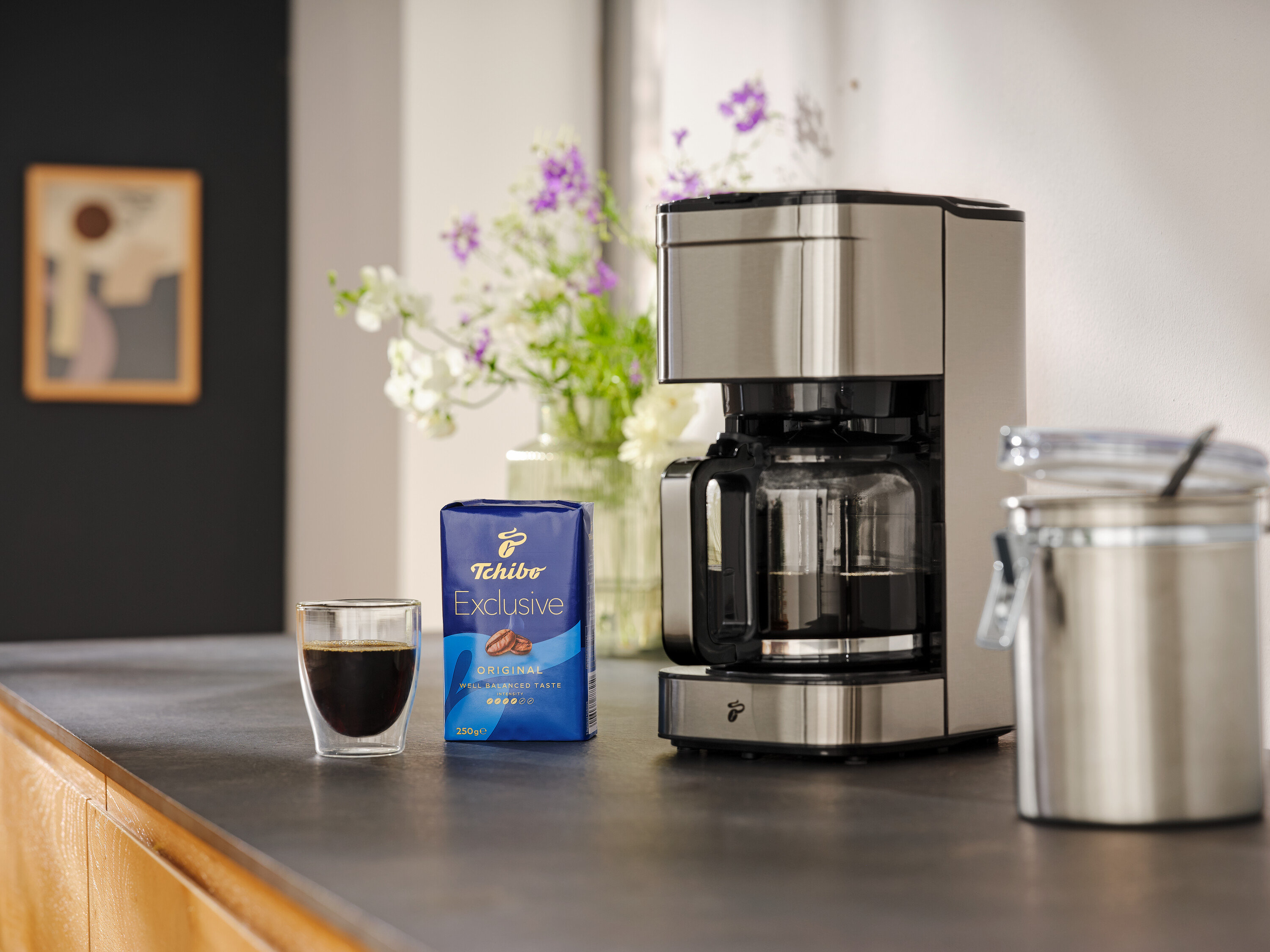 Na blacie kuchennym stoi ekspres do kawy, filiżanka kawy, paczka kawy Tchibo Exclusive i pojemnik do przechowywania.