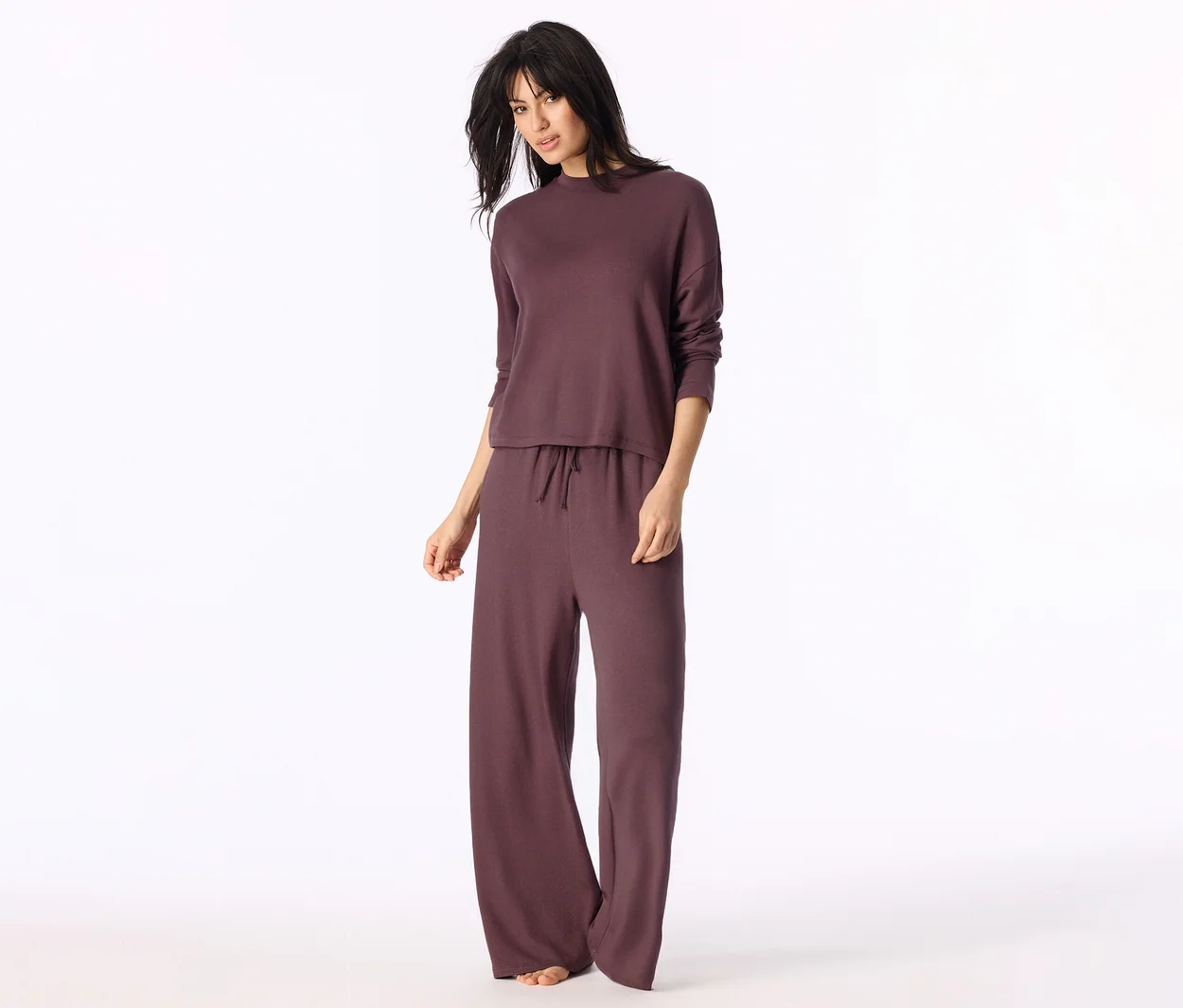 Kobieta w ciemnobordowym zestawie loungewear: bluzka z długim rękawem i szerokie spodnie ze sznurkiem. Stoi boso i patrzy w kamerę.