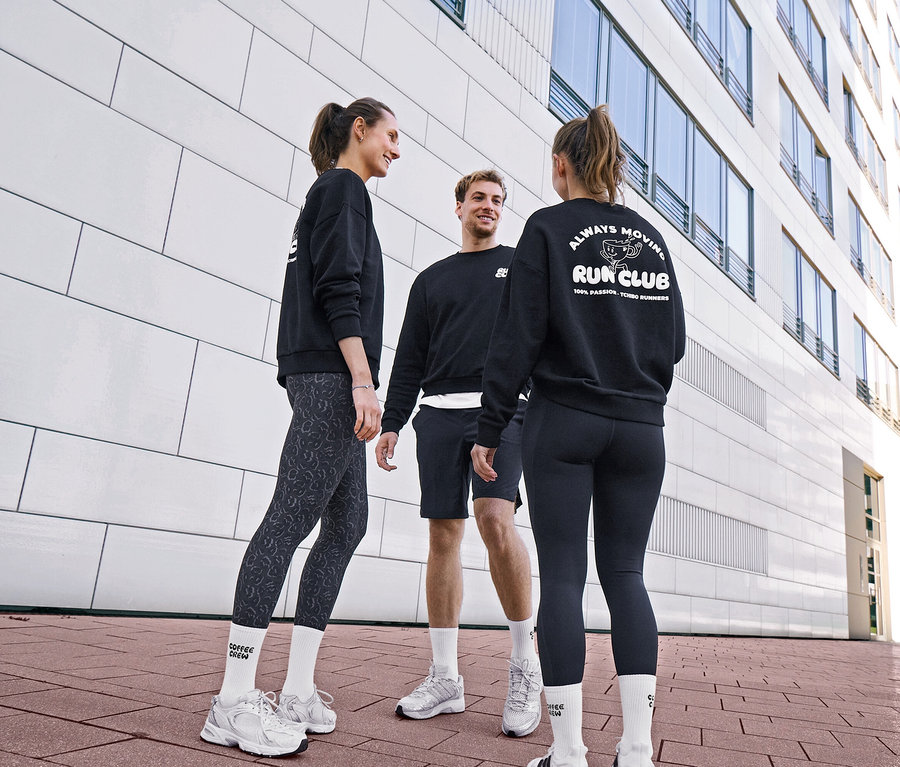 Trzy osoby w czarnych bluzach i strojach sportowych rozmawiają. Kobieta nosi szaro-czarne legginsy sportowe „ActivePower” i skarpety tenisowe Coffee Crew unisex. Mężczyzna nosi czarne szorty sportowe i białe skarpety. Inna kobieta nosi czarne legginsy sportowe „ActivePower” i bluzę z logo „RUN CLUB”.