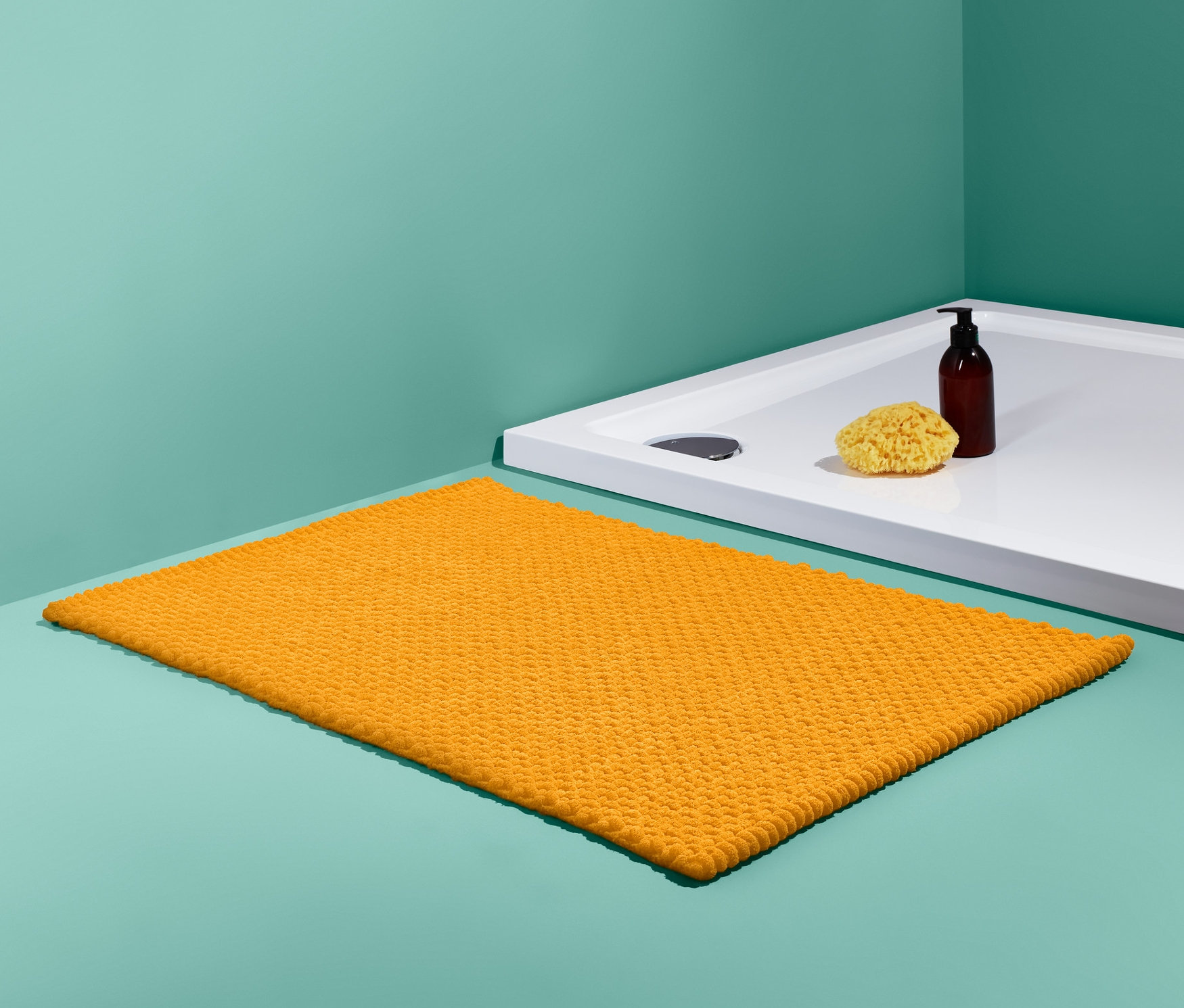 Pomarańczowa mata łazienkowa (ok. 50 x 80 cm) leży obok białego brodzika, na którym stoi brązowa butelka i żółta gąbka do mycia.