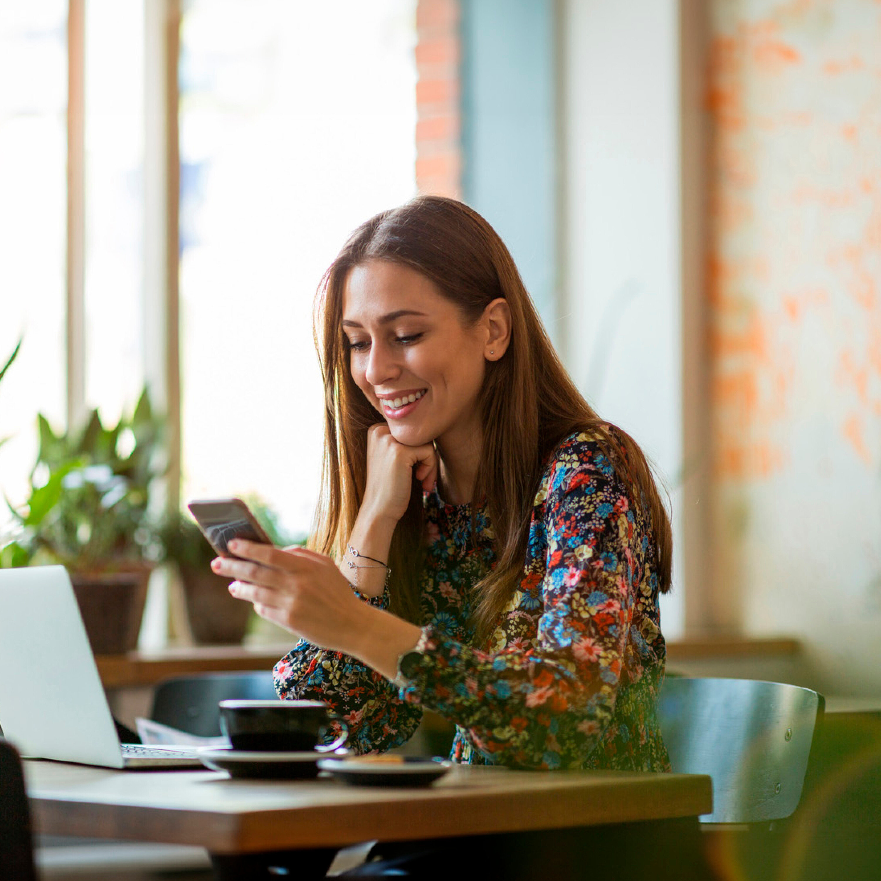 Uśmiechnięta kobieta patrzy w telefon komórkowy, siedząc przy stole z laptopem i filiżanką kawy.