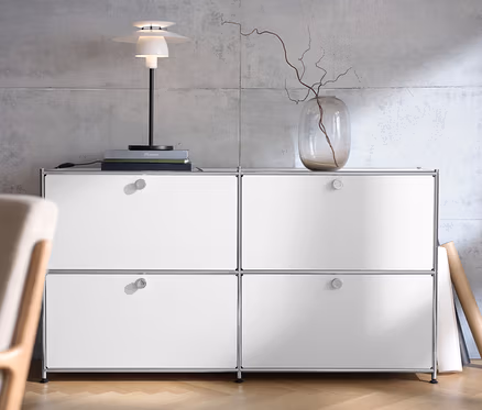 Biała szafka typu sideboard „CN3” z 4 klapami, na niej metalowa lampka stołowa i wazon z gałązkami.