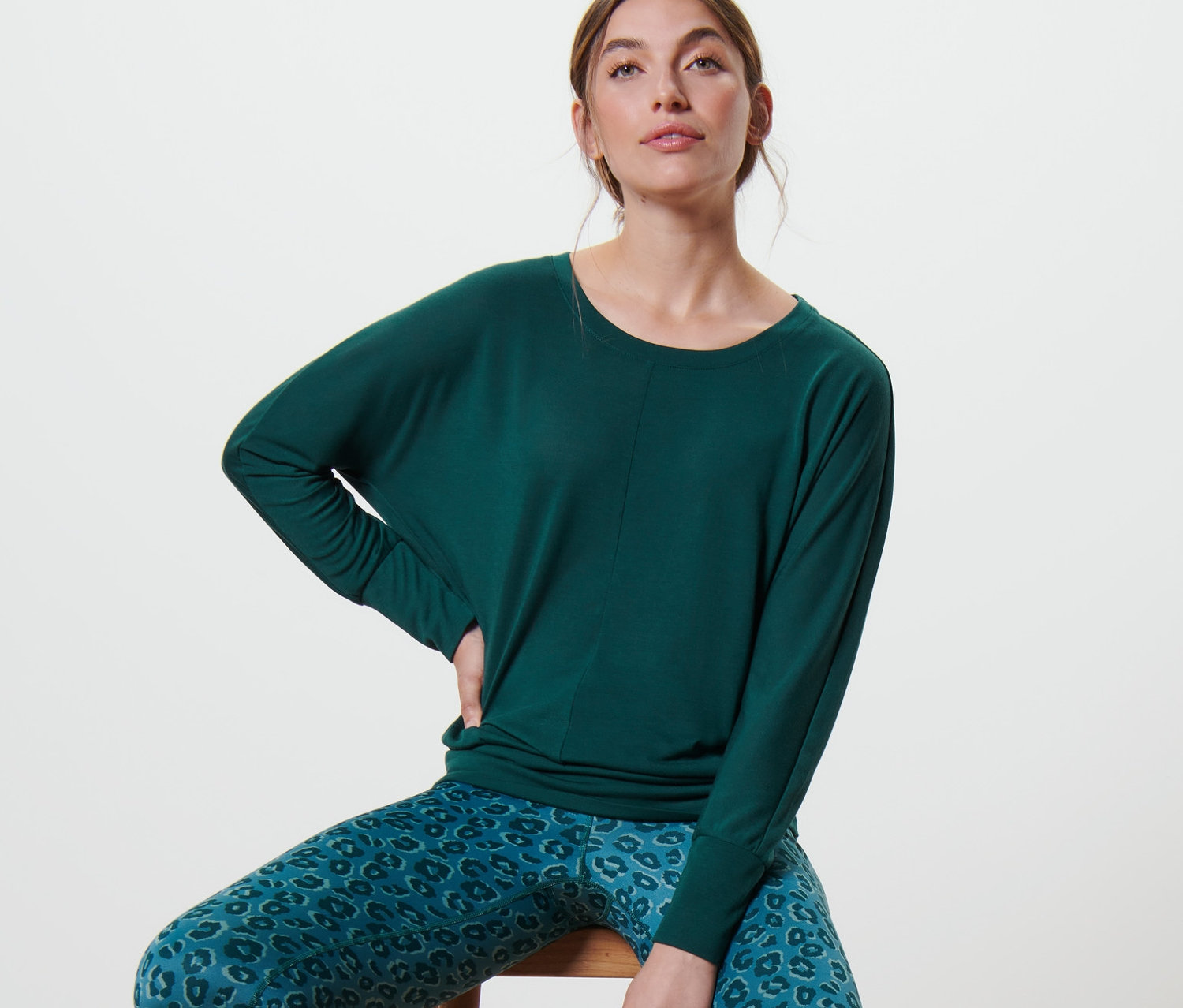 Kobieta siedzi na krześle, ma na sobie szmaragdową zieleń koszulki do jogi i treningu oraz niebiesko-zielone legginsy we wzór zwierzęcy. Pod krzesłem leży mata do ćwiczeń.