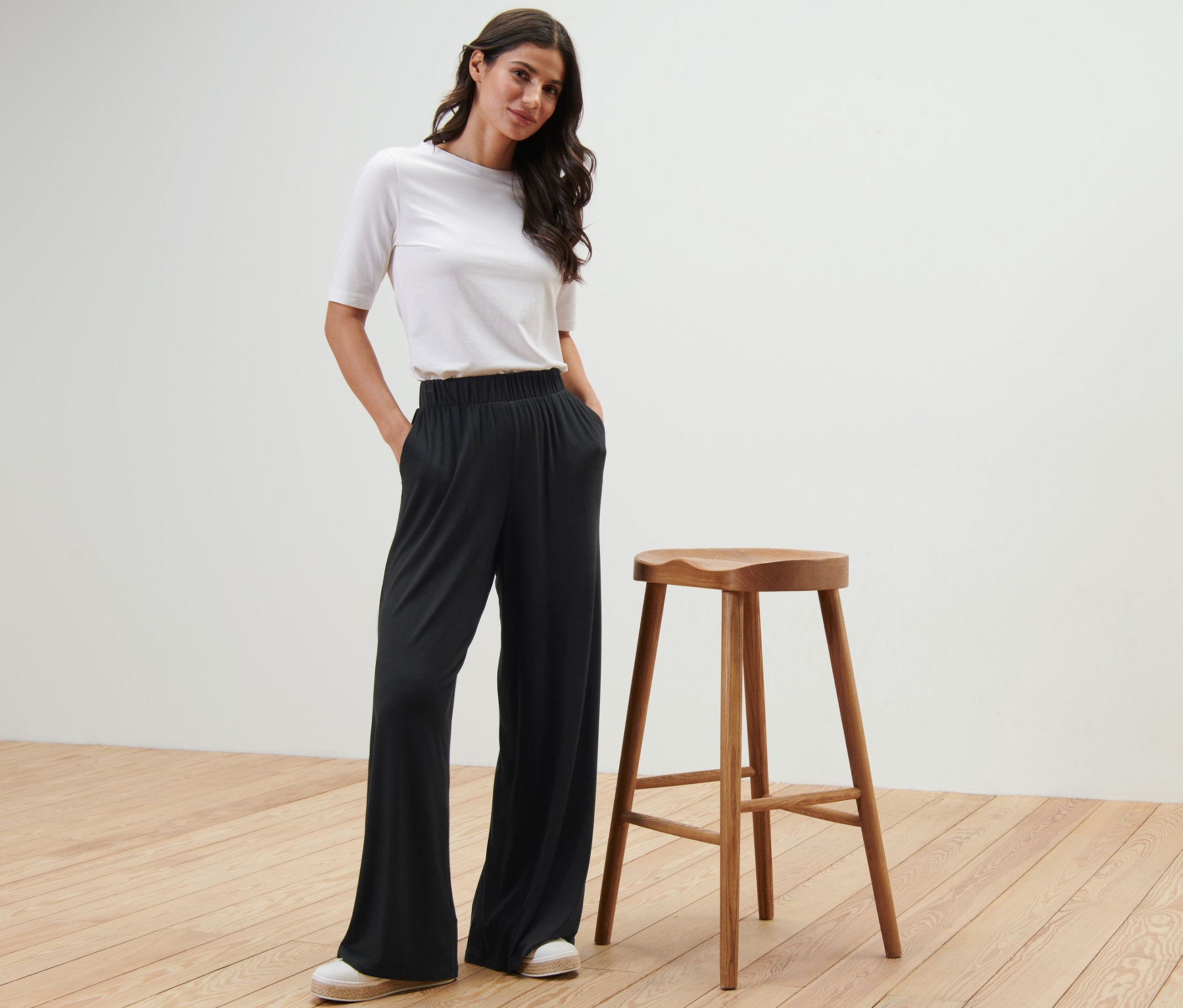 Kobieta pozuje w czarnych spodniach palazzo obok drewnianego stołka.