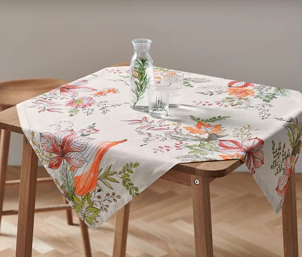 Na drewnianym stole leży obrus o wymiarach ok. 90 x 90 cm we wzory kwiatowe. Na obrusie stoi karafka z wodą i dwie szklanki.