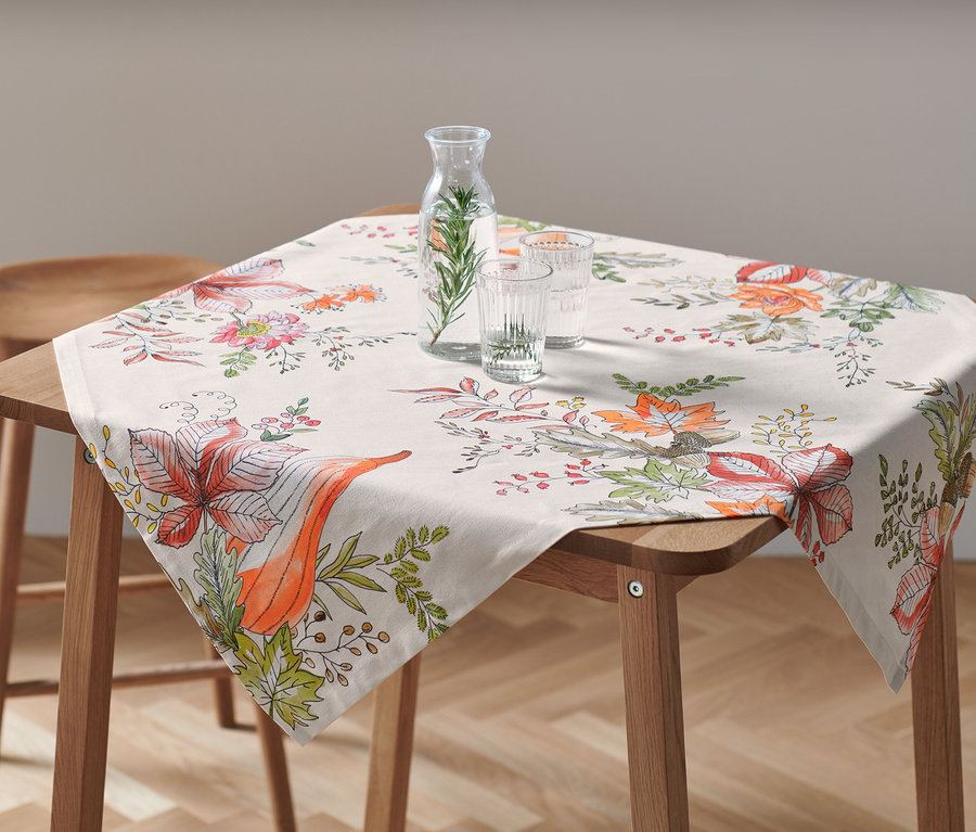 Na drewnianym stole leży obrus o wymiarach ok. 90 x 90 cm we wzory kwiatowe. Na obrusie stoi karafka z wodą i dwie szklanki.